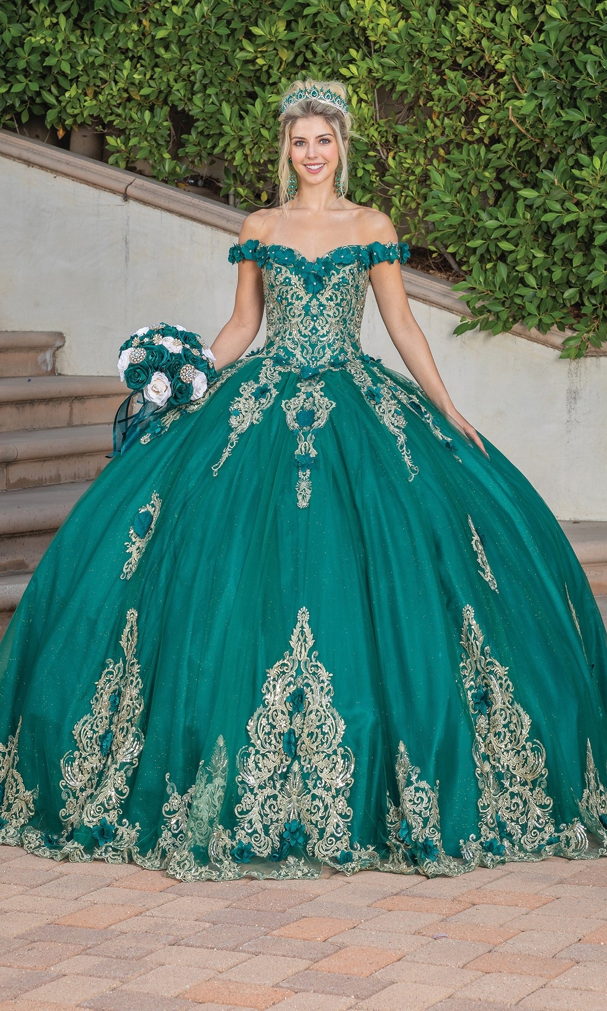 A young woman stands outdoors in the green and gold Quinceanera Dress 1739 By Dancing Queen, off the shoulder with lace embroidery, wearing a tiara and holding a bouquet.